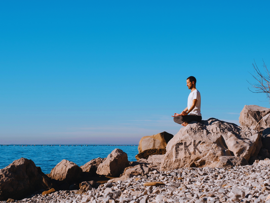 A man practicing the 4-7-8 breathwork technique sits cross-legged on a rocky shoreline, meditating under a bright blue sky with calm waters in the background, embodying mindfulness for anxiety relief